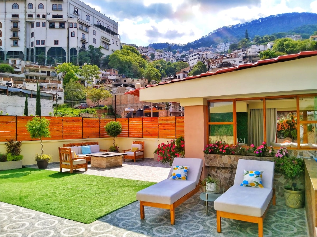 A spacious terrace features comfortable lounge chairs with colorful cushions, surrounded by greenery and vibrant flowers. A fire pit with seating is visible, providing a gathering space. The panoramic view of the hills and nearby buildings offers a tranquil backdrop for relaxation.