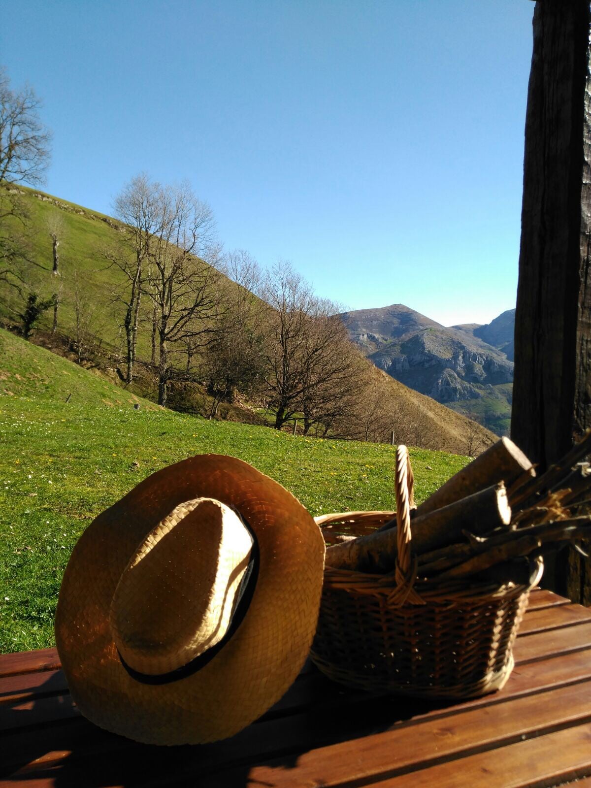 A natural landscape unfolds with lush green hills and a clear blue sky. A wooden table displays a straw hat next to a wicker basket filled with firewood, offering a rustic charm and a glimpse of the serene surroundings.