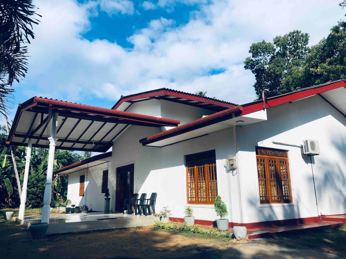 A modern single-story house is surrounded by greenery, featuring a red-tiled roof and white walls. The spacious front porch is furnished with black chairs, and large windows allow natural light to fill the interior. Lush trees are visible in the background.