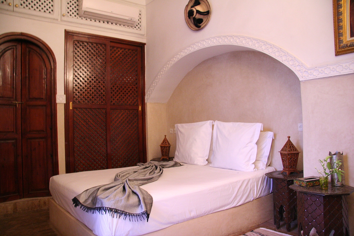A spacious bedroom is presented with a large, unmade bed featuring white pillows and a soft throw draped casually across the top. Decorative lanterns are placed on either side, and textured wall panels add depth to the serene atmosphere.