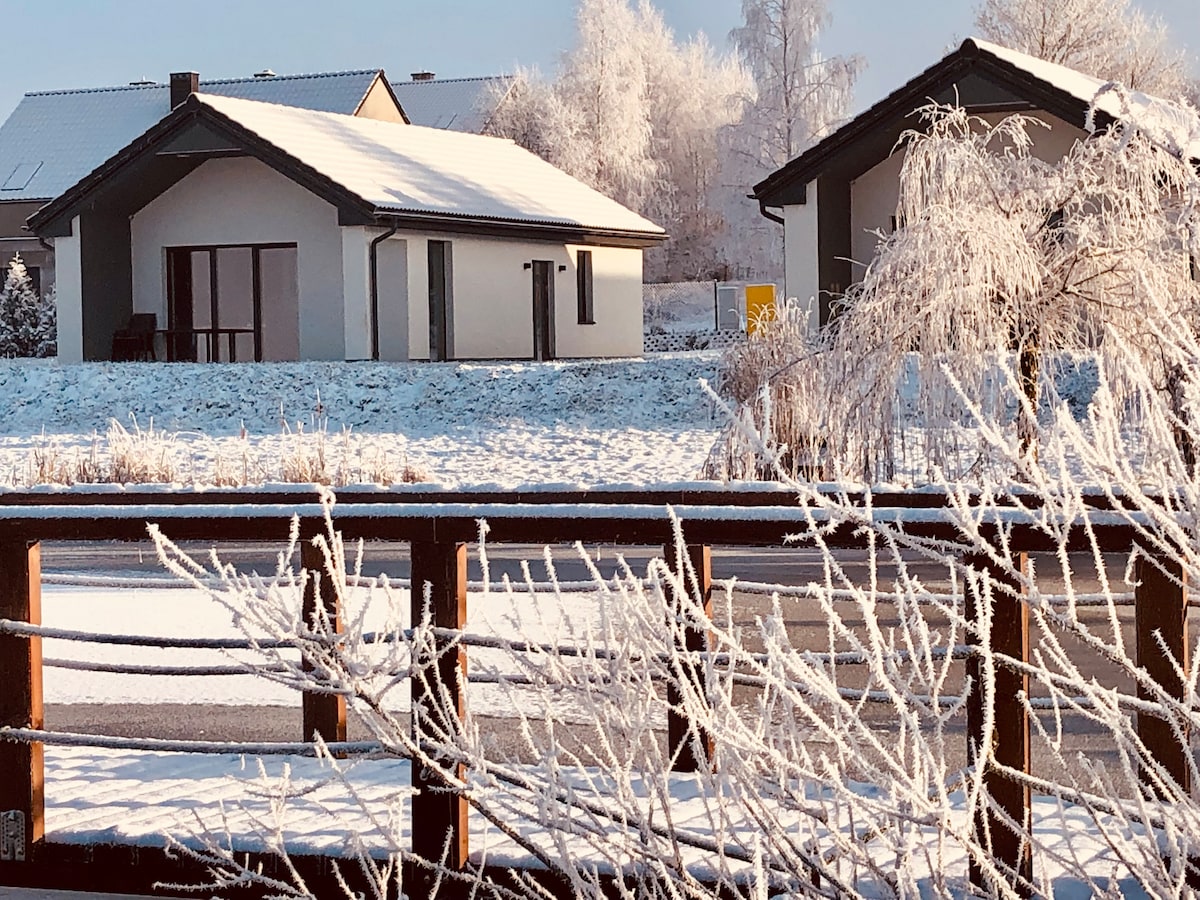 A winter scene features two modern houses with snow-covered roofs and frosted branches in the foreground. The landscape is blanketed in fresh snow, reflecting light. A wooden fence divides the scene, providing a rustic contrast to the contemporary architecture.