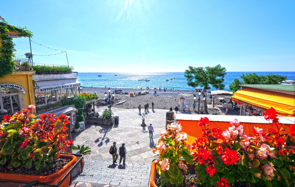 A vibrant outdoor scene is depicted, showcasing a beach area with a clear blue sky and calm sea. Colorful flower pots frame the view, while people stroll along the promenade. Restaurants and beach umbrellas can be seen in the background, inviting a lively atmosphere.