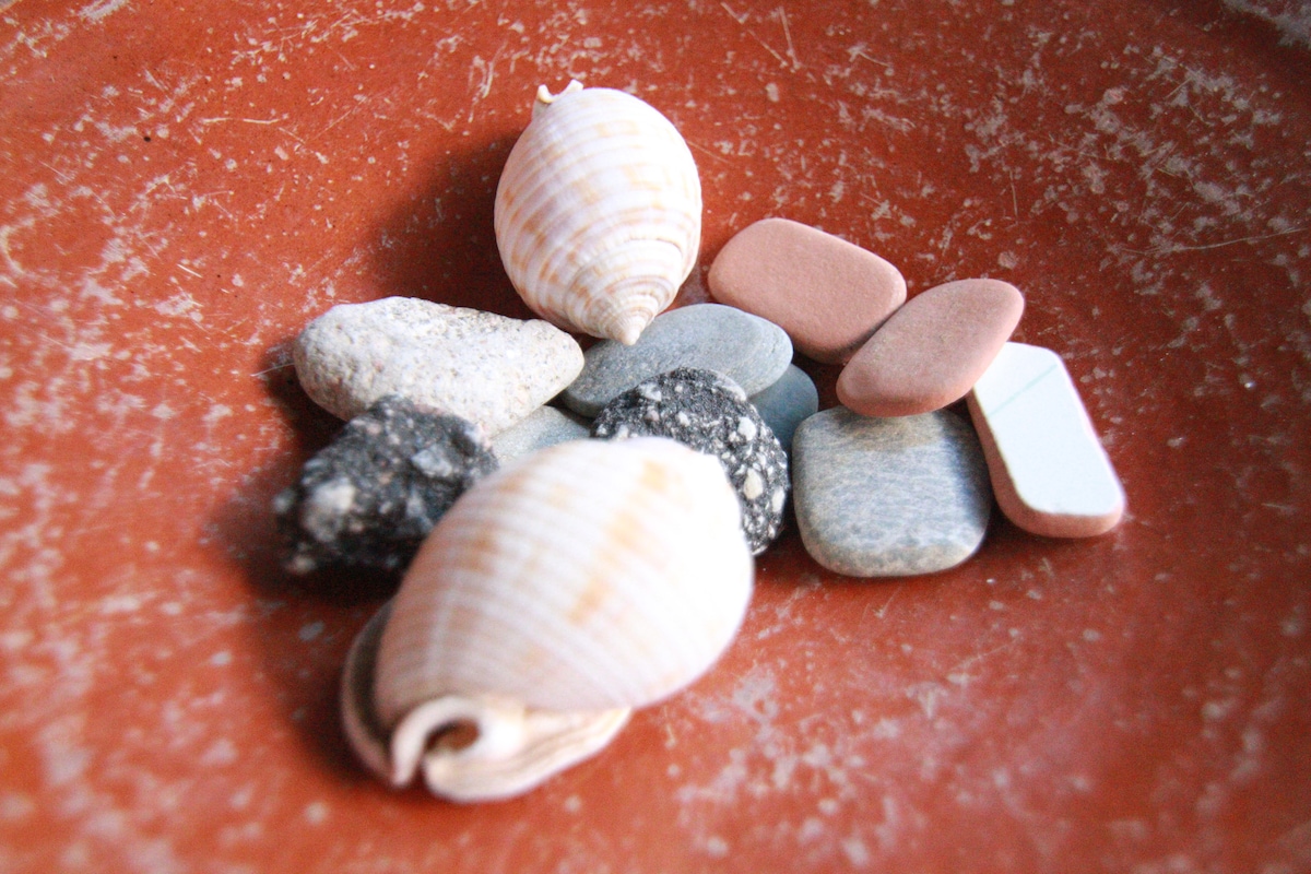 A shallow terracotta dish holds an assortment of natural items, including smooth pebbles and various shells. The colors of the items vary, featuring shades of grey, pink, and cream, creating a simple yet textured display.