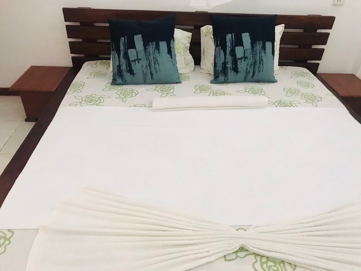 A spacious bed is centered in the room, adorned with vibrant green pillows and a white blanket. Two brown wooden side tables are positioned on either side of the bed, complementing the overall design. The bedding features a subtle floral pattern, enhancing the soft aesthetic.
