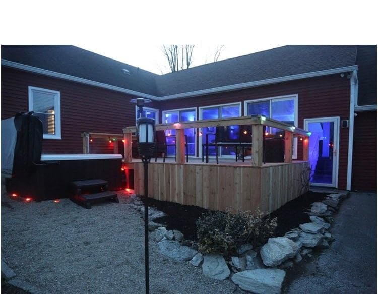 An outdoor deck is visible, surrounded by colorful lighting. A hot tub is positioned on one side, with seating and a table that allows for relaxation. The area is landscaped with gravel and small stones, creating a welcoming atmosphere for gatherings.