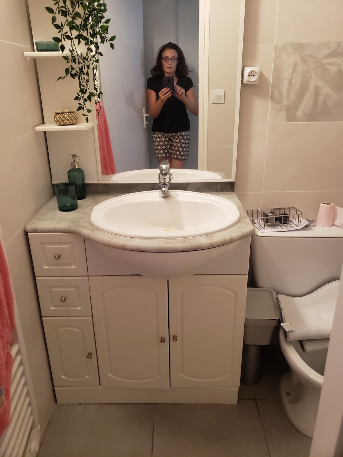 The bathroom features a white vanity with a circular sink and stone countertop, complemented by a mirror above. Shelves display decorative glass bottles, while a towel hangs on a nearby rail. A compact toilet and a small waste bin are visible in the corner.