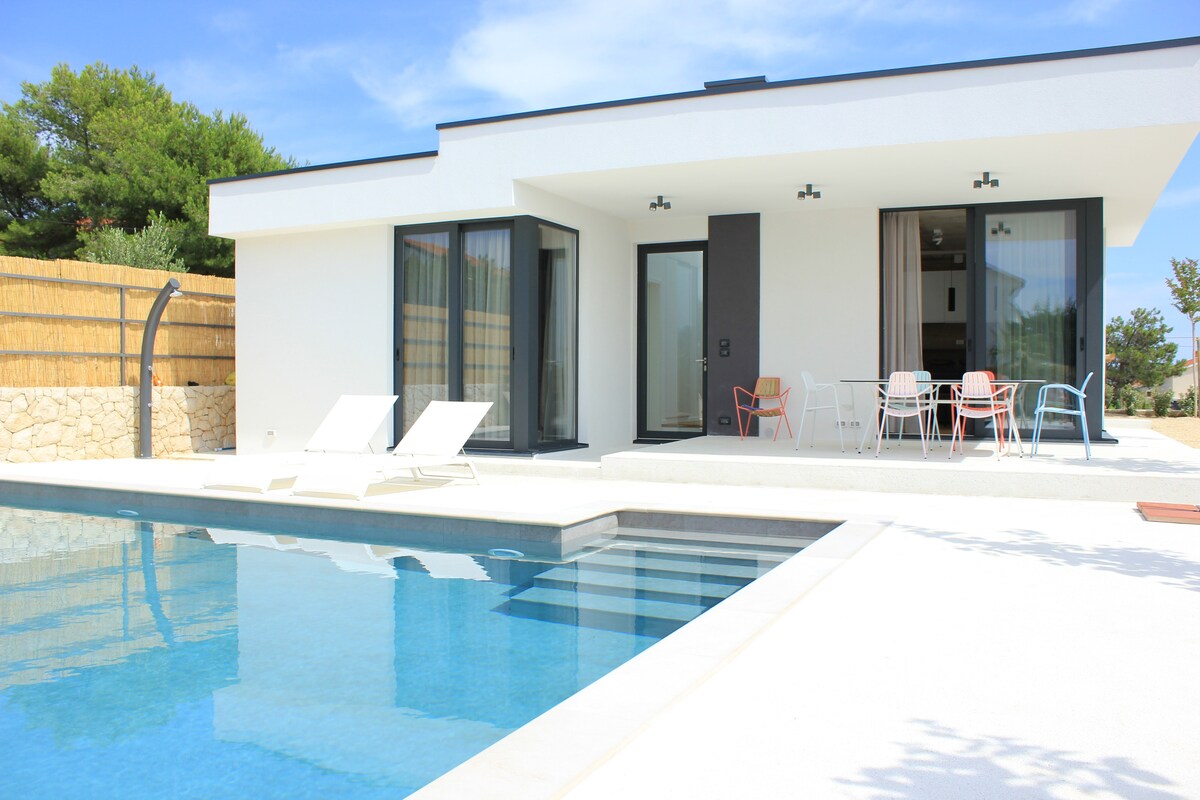 The outdoor area features a spacious terrace with a large swimming pool reflecting the clear sky. Sun loungers are positioned near the water, while a dining area with colorful chairs is visible in the background. The house's modern architecture is complemented by large glass doors.