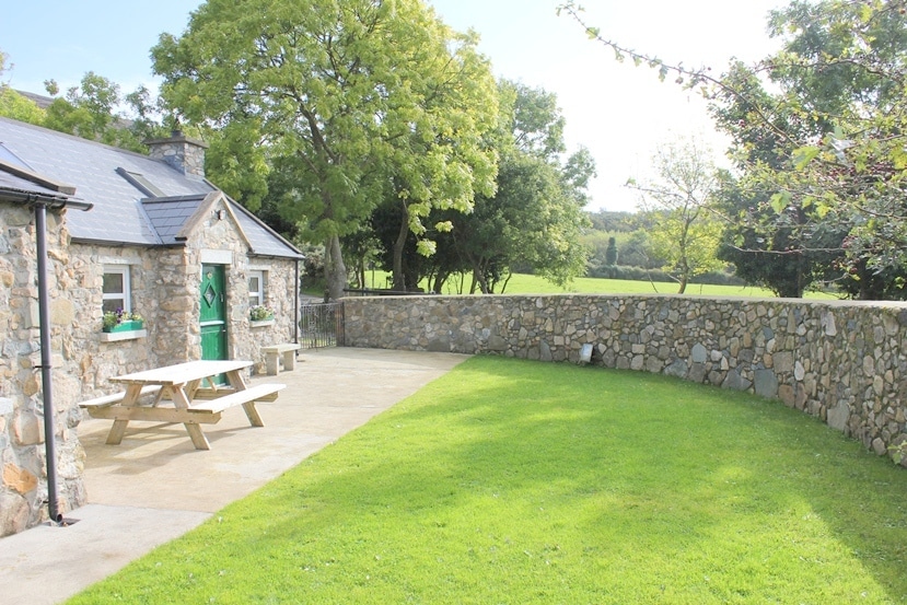 A charming stone cottage is highlighted in the image, featuring a green door and surrounded by lush grass. An outdoor seating area with a picnic table is situated on a smooth concrete patio. The surrounding area includes a stone wall and vibrant trees, enhancing the peaceful setting.