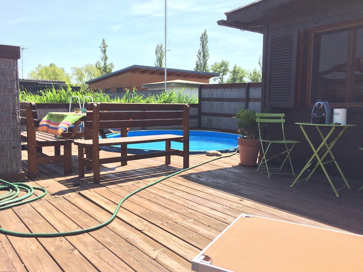 The outdoor area features a wooden deck surrounding a private pool, with two green chairs and a small table for relaxation. A bench extends along the edge of the pool, surrounded by greenery, creating a space for leisure and enjoyment. Sunlight illuminates the setting.