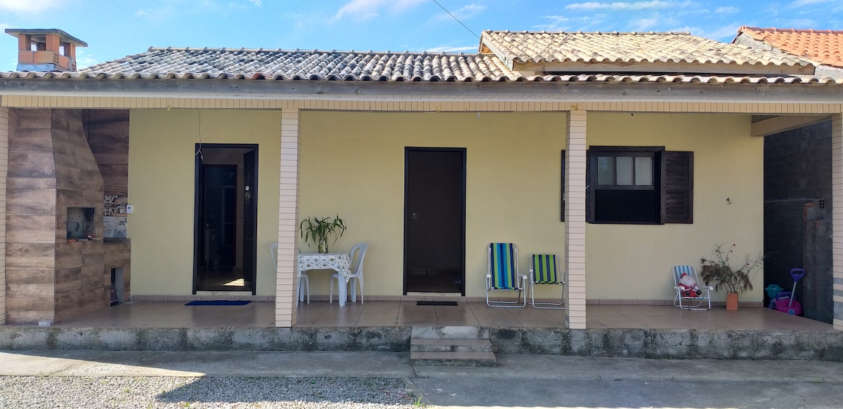 A single-story house is shown with a tiled roof and welcoming entrance. A small set of steps leads to a porch with two colorful chairs and a table. Windows are present on either side of the door, allowing natural light to enter the space.