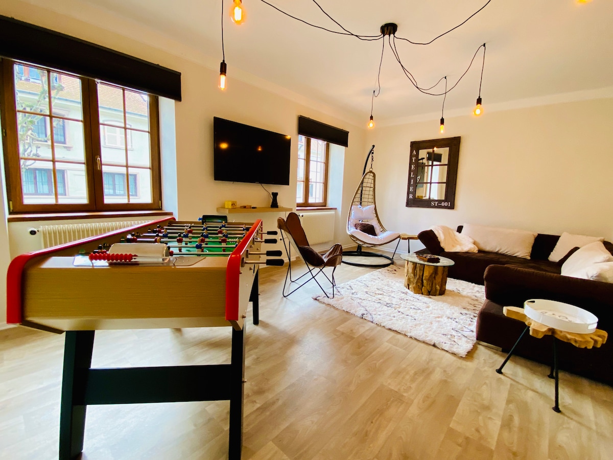 A bright living area showcases a foosball table and a comfortable seating arrangement. Wall-mounted entertainment is paired with natural light from large windows. Unique lighting fixtures enhance the ambiance, while the cozy decor includes a wool rug and wooden accents, contributing to a welcoming atmosphere.
