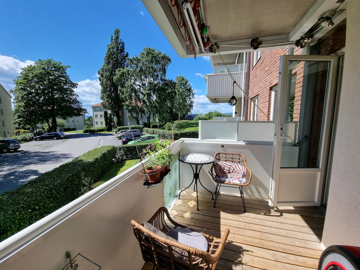 A balcony space features wooden flooring and a small round table accompanied by two chairs. Potted plants add touches of greenery. A view of grassy areas and trees is visible beyond the railing, showcasing a calm neighborhood atmosphere.
