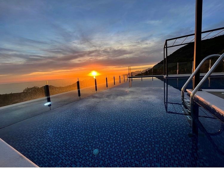 A rooftop pool is featured with a stunning view of the sunset. The water reflects the vibrant colors of the sky, and glass railings surround the pool area, enhancing the serene atmosphere. The distant hills create a peaceful backdrop.