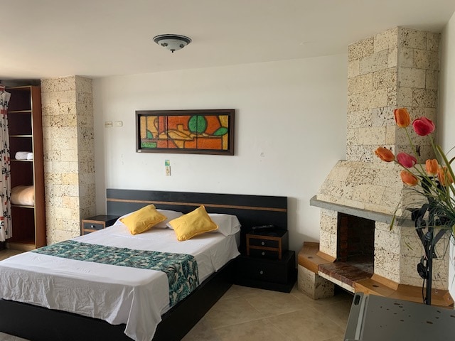 A spacious room features a large bed adorned with two yellow pillows and a decorative green runner. A stained glass artwork is displayed above the headboard. An elegant fireplace and a vase with vibrant flowers add a touch of warmth to the minimalist design.