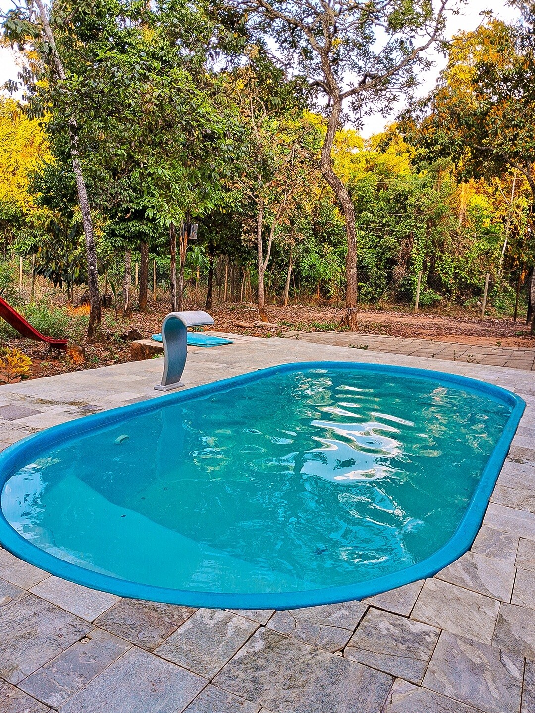 The pool area features a rectangular, clear blue swimming pool surrounded by a stone patio. Lush green trees and vegetation add a natural backdrop, providing a secluded atmosphere. A slide is visible, enhancing the recreational space.