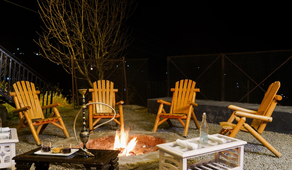 A cozy outdoor bonfire area features four rustic wooden chairs arranged around a fire pit. The setting is illuminated by the flickering flames, with a table displaying snacks and drinks nearby, surrounded by a serene night atmosphere.