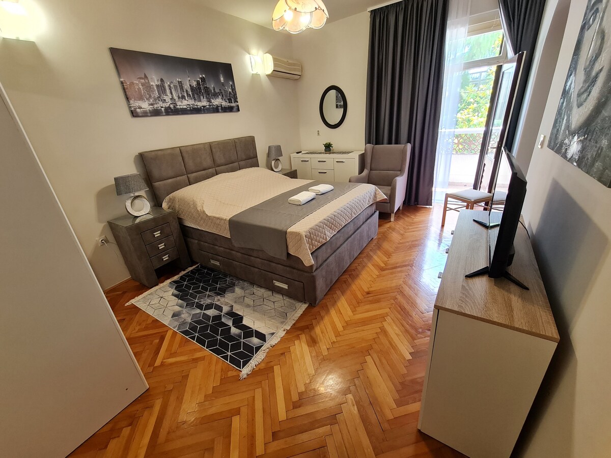 A spacious studio apartment features a comfortable bed with a neutral bedding arrangement. A large piece of wall art depicting a city skyline adds visual interest. Natural light is accessible through a window covered by flowing curtains. A dresser, television, and a patterned rug complete the inviting interior.