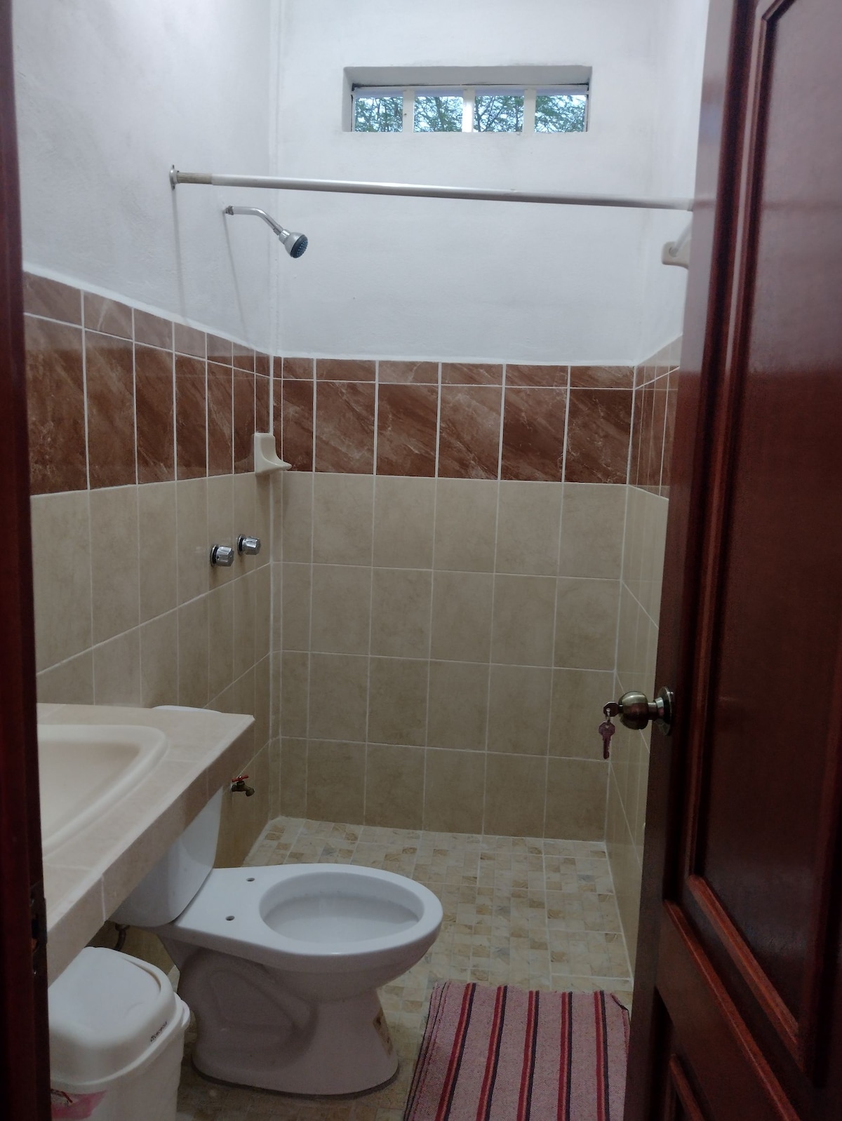 A well-lit bathroom is visible, featuring tiled walls with a combination of light and dark hues. A showerhead is mounted above a neutral-colored bathtub, and a toilet is positioned beside a sink with a countertop. A small window allows natural light to enter.