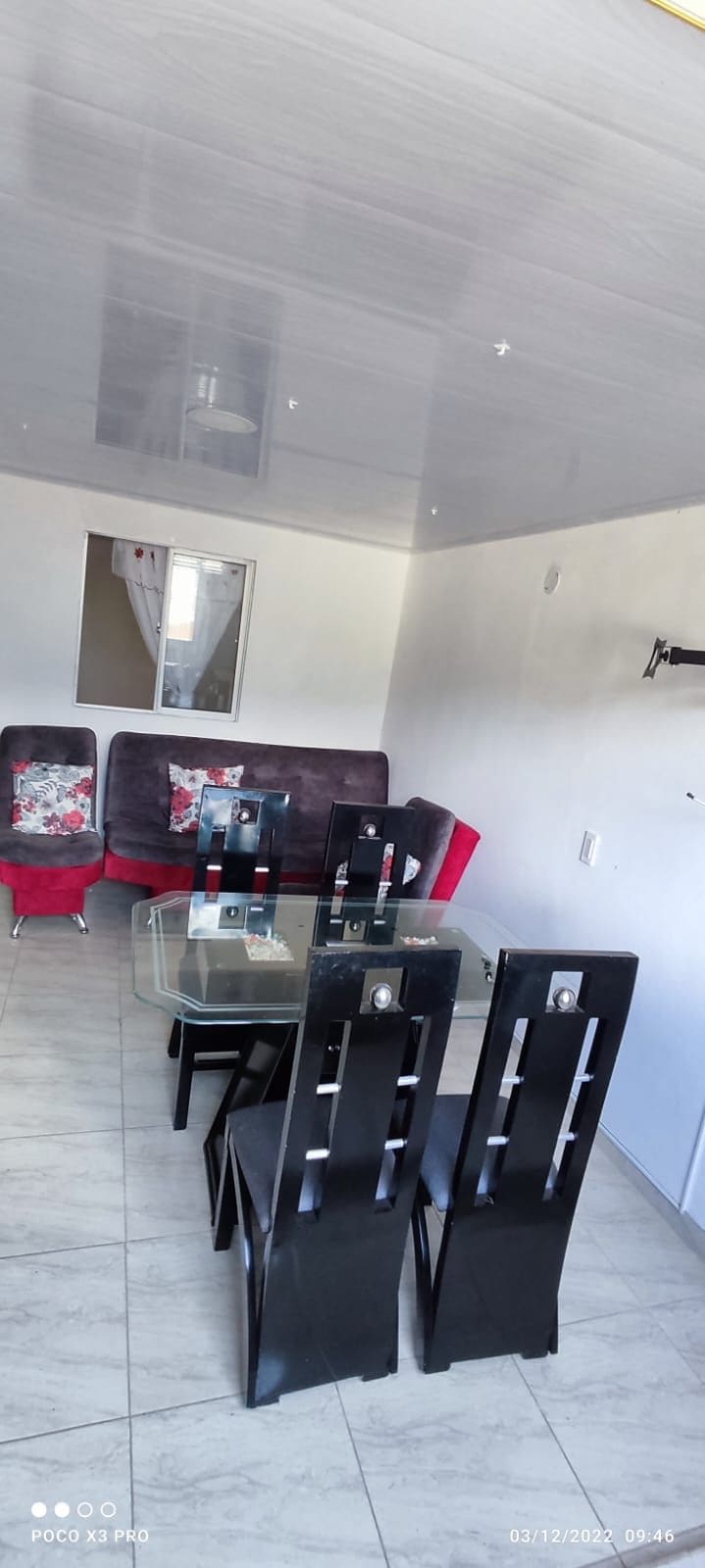 A dining area is presented with a glass table surrounded by black chairs. A comfortable sofa with decorative pillows is visible in the background. Light-colored tiles and a small window offer natural illumination throughout the space.