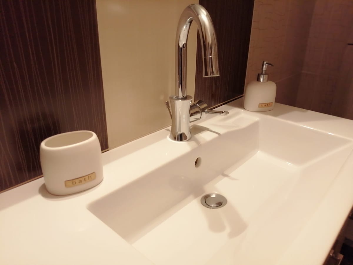 A modern bathroom sink is showcased, featuring a sleek faucet and a white basin. Two white containers with the label 'bath' are positioned beside the sink. The background displays elegant dark wood paneling, enhancing the contemporary aesthetic.
