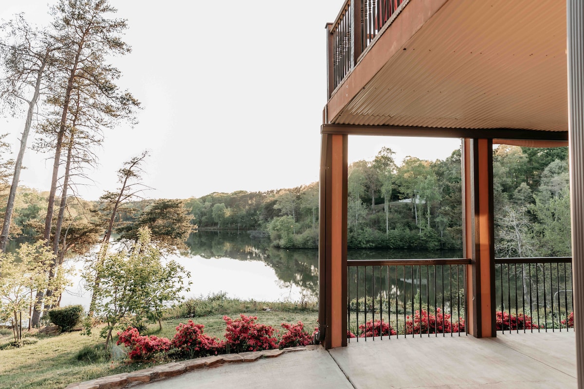 A private balcony offers views of Blalock Lake, framed by lush greenery and vibrant blooming flowers. The calm water reflects the surrounding trees, creating a peaceful setting. The balcony features a metal railing, providing a safe space to enjoy the scenic landscape.