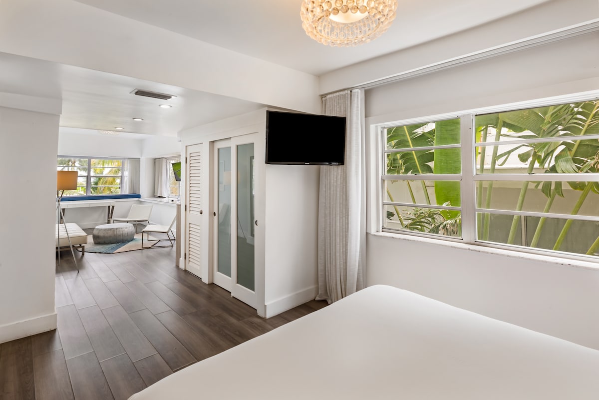 A bright interior space features a spacious bed in the foreground and a flat-screen TV mounted on a wall. Windows allow natural light to fill the room, revealing views of lush greenery outside. The warm wood flooring adds to the inviting atmosphere.