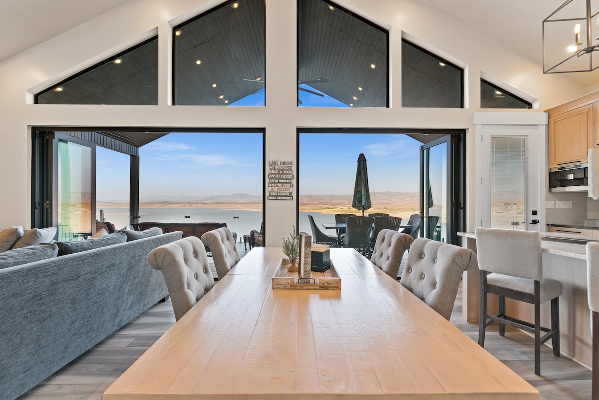 A spacious dining area is centered by a large wooden table, surrounded by upholstered chairs. Large glass doors open to a breathtaking lake view, allowing natural light to fill the room. Comfortable seating arrangements provide a cozy atmosphere for gatherings.