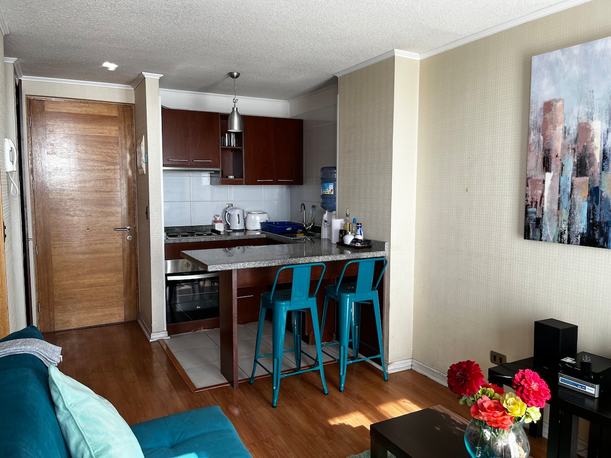 A cozy kitchen area features wooden cabinetry and a granite countertop. An array of kitchen appliances is present, including a kettle and stove. Two blue bar stools complement a small dining space, while a colorful painting adds a pop of interest to the wall.