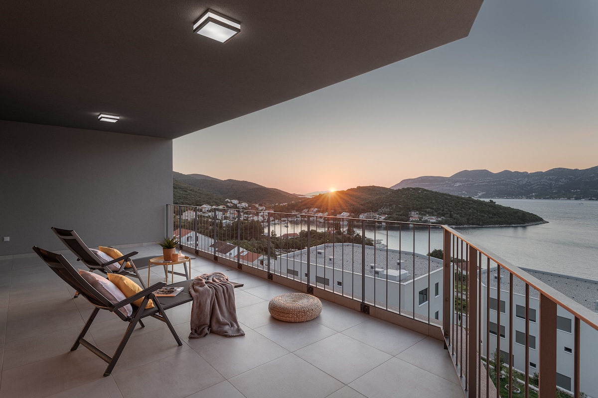 A private balcony is showcased with two loungers, facing the breathtaking sunset over the calm waters of the bay. A soft blanket and a floor cushion are placed nearby, while the distant hills provide a serene backdrop against the fading light.