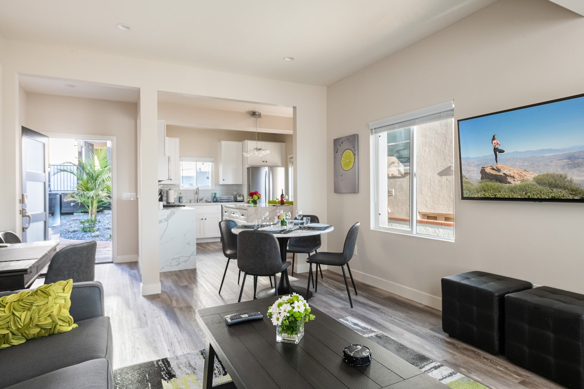 An open floor plan connects the living area to the kitchen, with a dining table set for four. A large window allows natural light to illuminate the space, while a modern TV is mounted on the wall. Comfortable seating is arranged nearby.