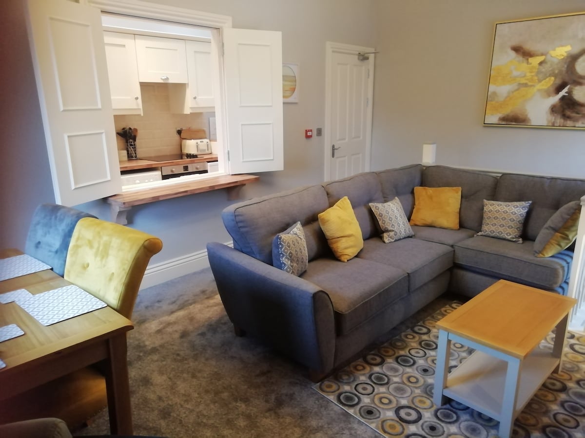 A spacious living area features a comfortable gray sectional sofa adorned with yellow accent pillows. Nearby, a wooden dining table with two chairs is complemented by a decorative area rug. A well-equipped kitchen is partially visible through the open doorway.