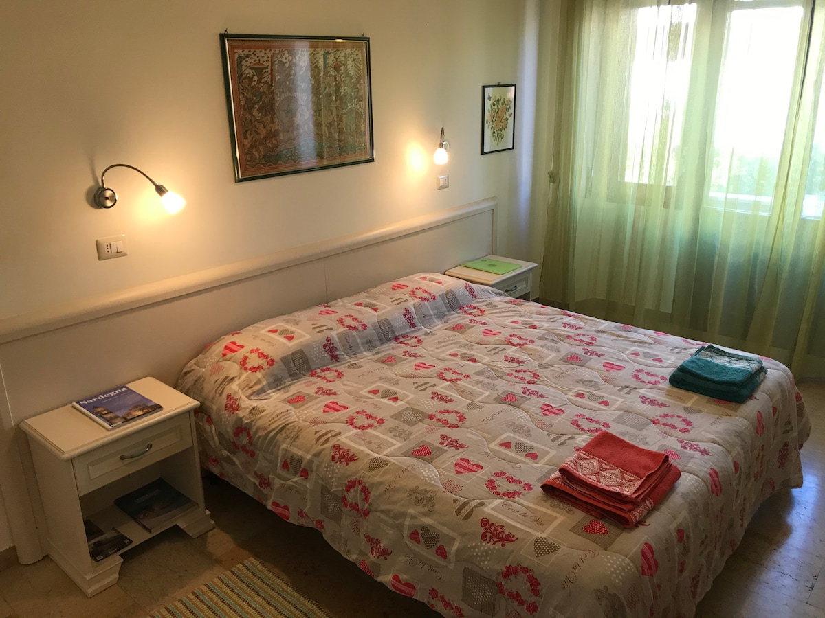 A spacious bedroom is presented with floral-printed bedding on a large bed. Two bedside tables are positioned alongside the bed, each with wall-mounted reading lights. Natural light enters through large windows adorned with sheer, green curtains, enhancing the calming atmosphere.