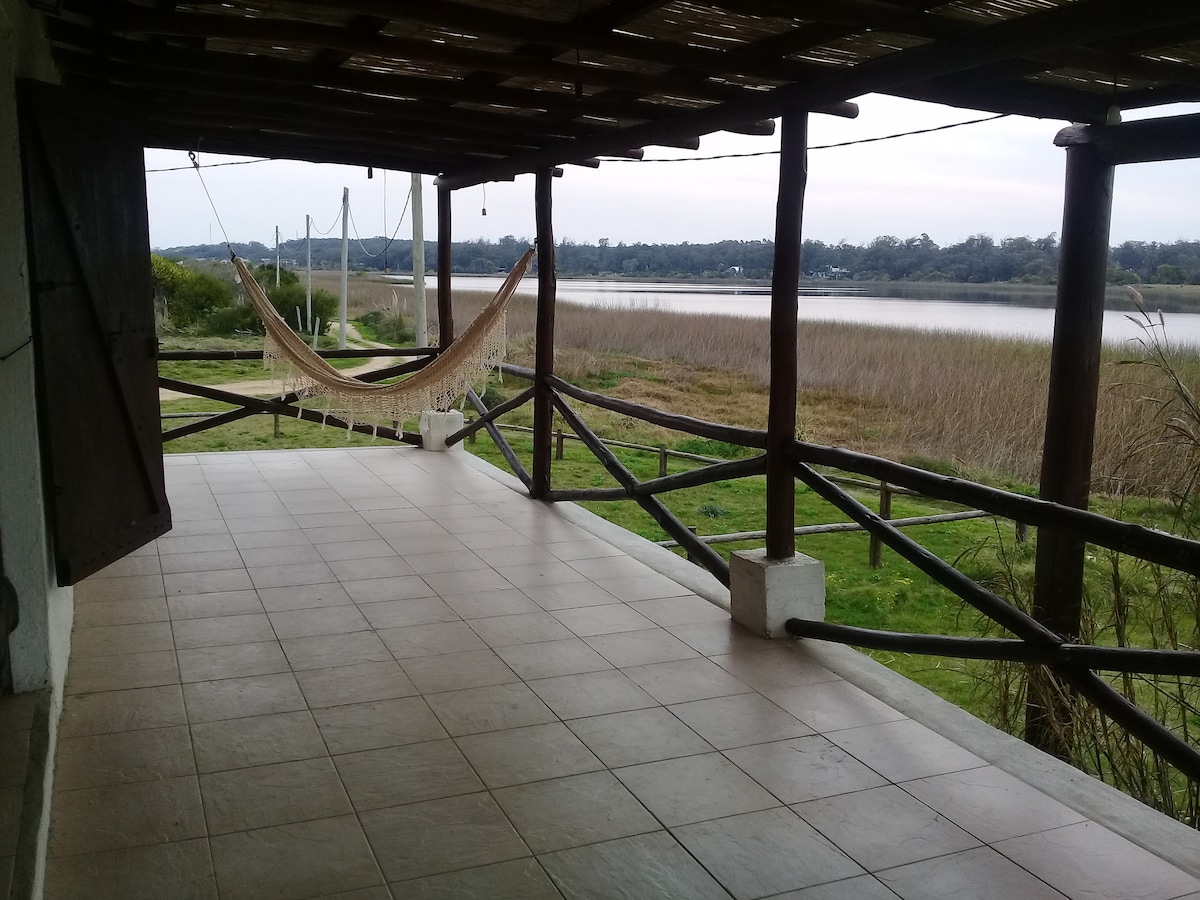 A spacious terrace features a woven hammock suspended between wooden beams, overlooking a scenic water view. The area is framed by rustic wooden railings and offers a tiled floor that enhances the open, airy atmosphere, making it inviting for relaxation.