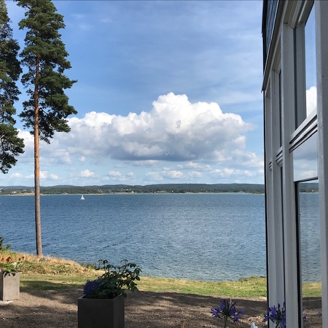 A wide view of Hakefjorden is captured, showcasing calm waters reflecting the sky. Lush greenery and trees frame the scene, while a sailboat can be seen in the distance. The modern structure with large windows allows for an unobstructed view of the tranquil landscape.