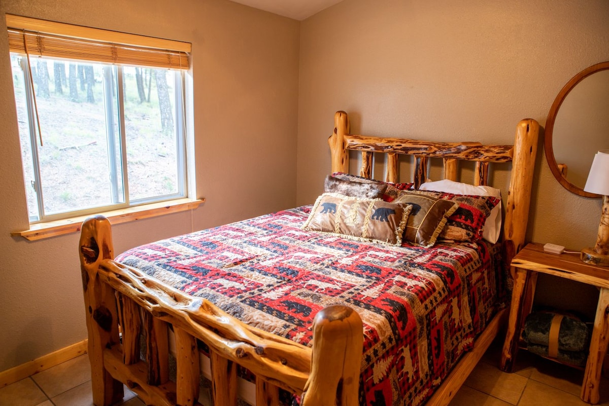 A cozy bedroom features a queen bed framed with rustic wooden posts and adorned with a patterned quilt. A round mirror and a nightstand complement the room, with natural light illuminating the space through a nearby window.