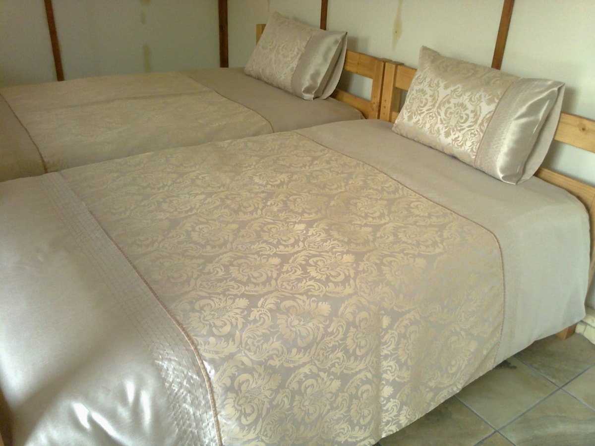 Two spacious beds are arranged in the room, each adorned with elegant beige bed coverings and matching decorative pillows. The wooden bed frames add a natural touch, complementing the tiled floor and neutral wall backdrop.