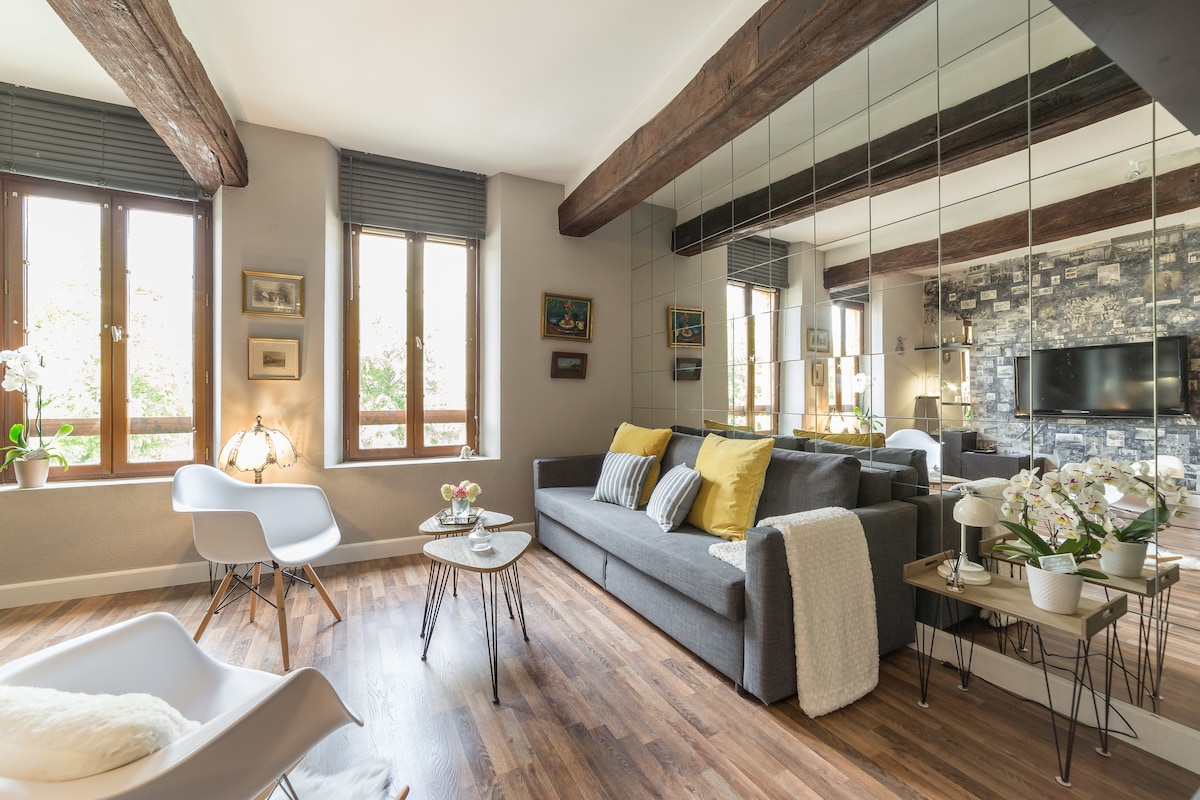 A cozy living area is highlighted by natural light streaming through multiple windows. A modern gray sofa is accented with yellow pillows, facing a wall-mounted television. Two small tables with floral arrangements are present, along with white chairs providing additional seating.