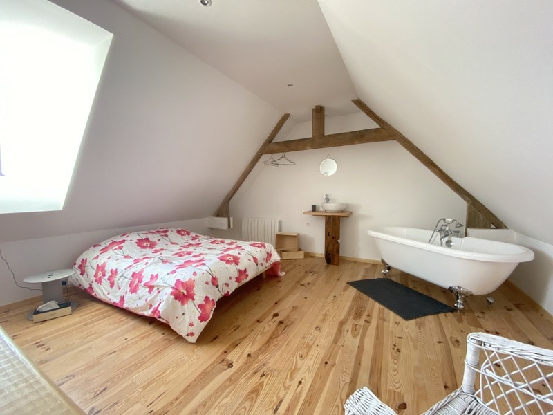 A serene bedroom is presented with a spacious bed adorned in floral bedding. A vintage-style clawfoot bathtub sits beside the bed. Natural light filters through a sloped window, highlighting the wooden beams and polished floor. A small bedside table and mirror complete the arrangement.