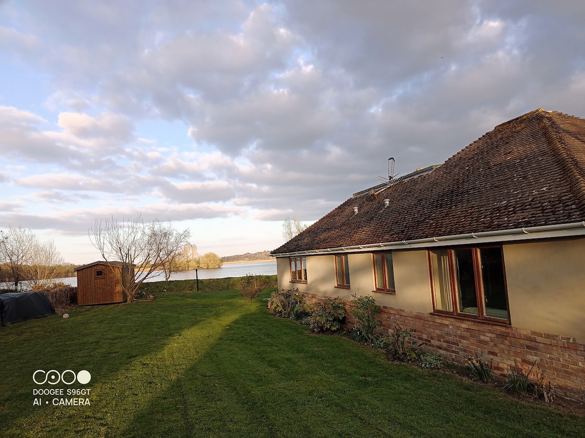 The exterior of the bungalow is depicted, showcasing a grassy area leading to the lake. Large windows allow for natural light, while nearby trees and shrubs enhance the natural setting. A small shed is visible, set against a backdrop of calm water and a partly cloudy sky.
