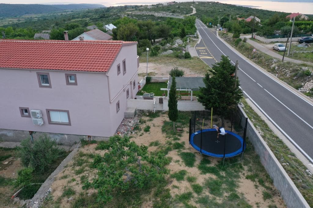 An aerial view of the property showcases a newly built apartment in a rural setting, surrounded by greenery. A large trampoline is visible in the yard, offering a space for recreation. The nearby road is lined with trees, and the landscape extends into the distance.
