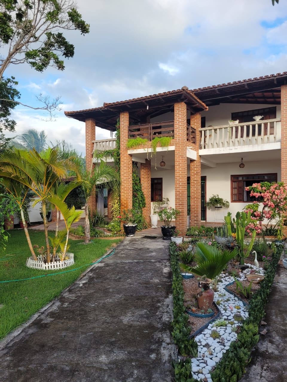The exterior of a two-story home is presented, featuring brick columns and a spacious balcony. Lush greenery surrounds the property, including palms and flowering plants. A well-maintained garden path composed of stones and decorative elements leads to the entrance, enhancing the natural setting.