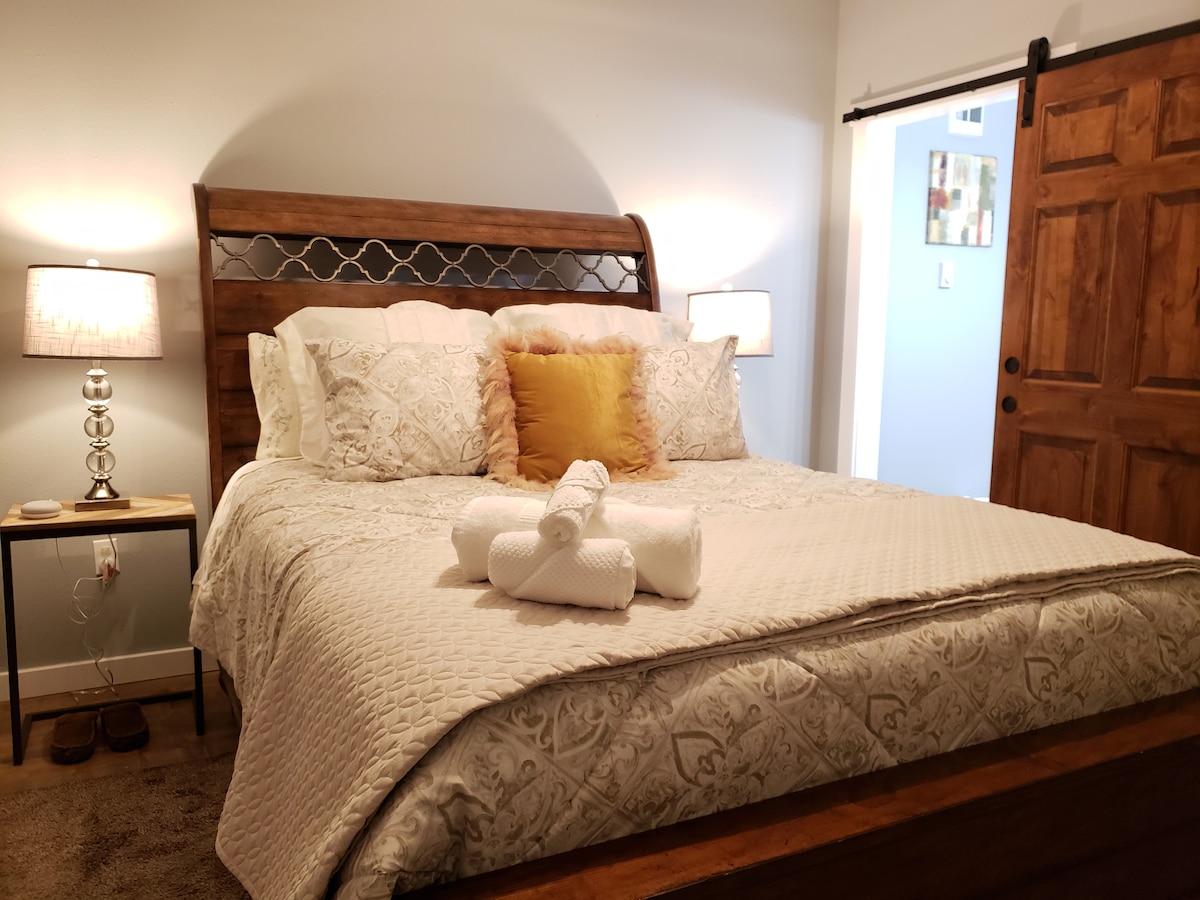 A spacious bed with a decorative headboard is centered in the room, featuring soft bedding and a yellow accent pillow. Nightstands with lamps flank the bed, providing light. A set of rolled towels is neatly arranged at the foot of the bed, enhancing the cozy ambiance.