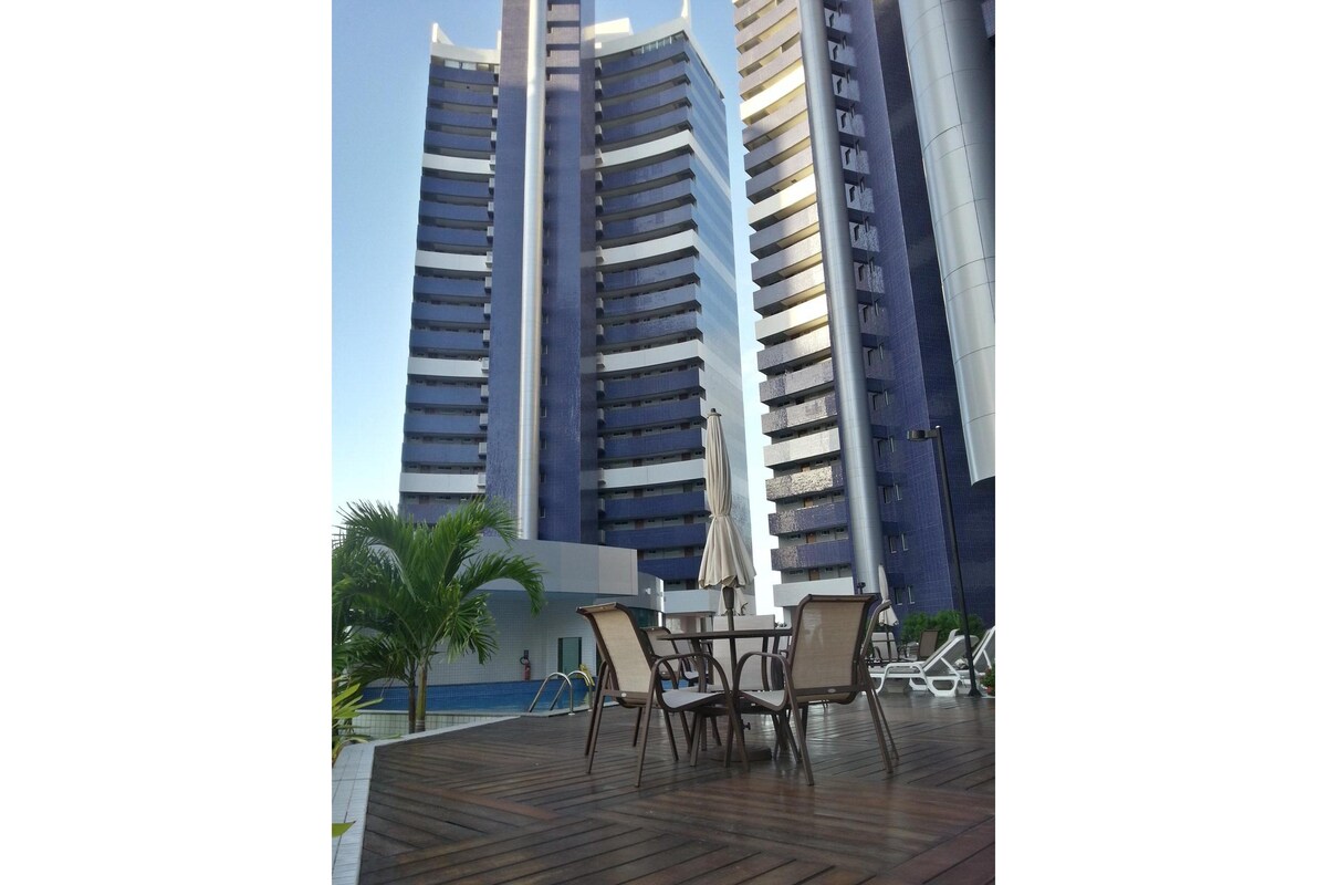 The outdoor area features a wooden deck with a dining table and chairs. A large umbrella provides shade, surrounded by tropical plants. In the background, the modern condominium towers rise, showcasing multiple levels and sleek architectural design. A swimming pool is visible to the left.