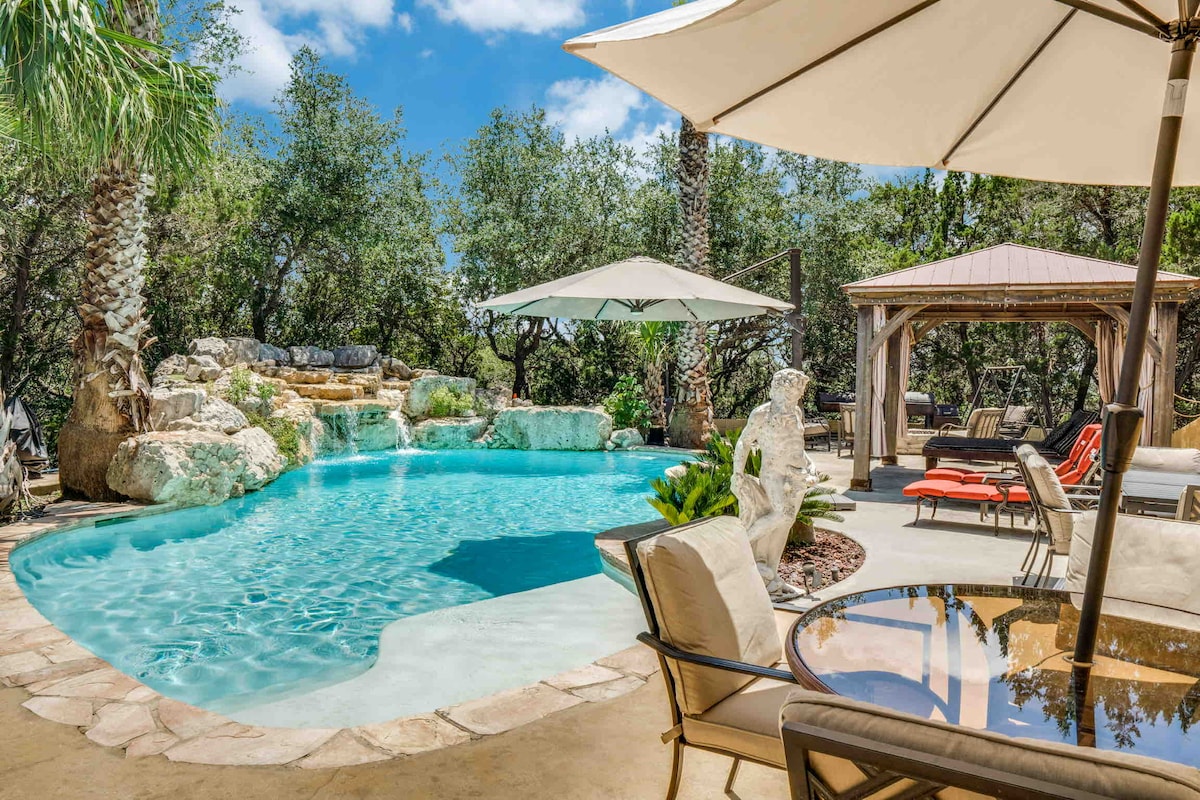 The outdoor pool area features a large swimming pool with a natural rock waterfall, surrounded by lush greenery. Comfortable lounge chairs are placed nearby, and a shaded seating area is provided under a spacious umbrella, inviting relaxation in a serene setting.