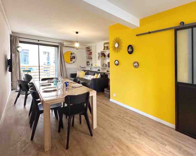 A bright dining area features a wooden table surrounded by black chairs, accommodating six. A sunny yellow wall adds warmth to the space, while a comfortable sofa with decorative cushions is visible in the background. Natural light enters through large glass doors, enhancing the inviting atmosphere.