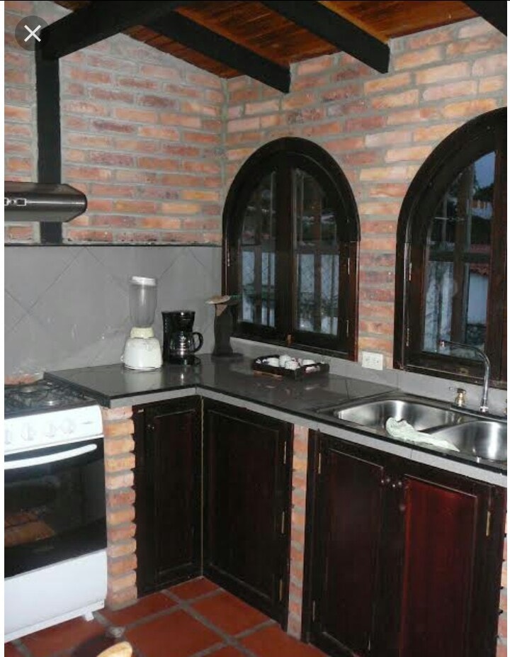 An updated kitchen features dark wooden cabinetry and grey countertops. A gas stove and an oven are present, alongside a blender and a coffee maker. Large arched windows allow natural light to fill the space, highlighting the brick wall and tile flooring.