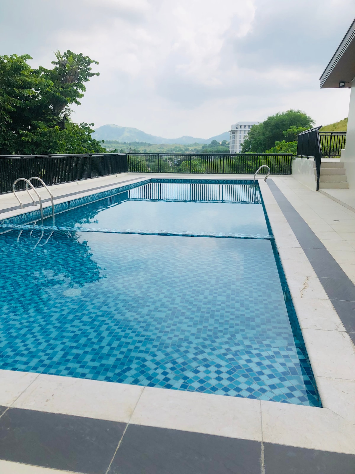 A tranquil outdoor pool is displayed, featuring clear blue water and a mosaic tile pattern. Surrounding greenery and distant hills can be seen, while a sleek railing runs along the poolside, providing a serene environment for relaxation.