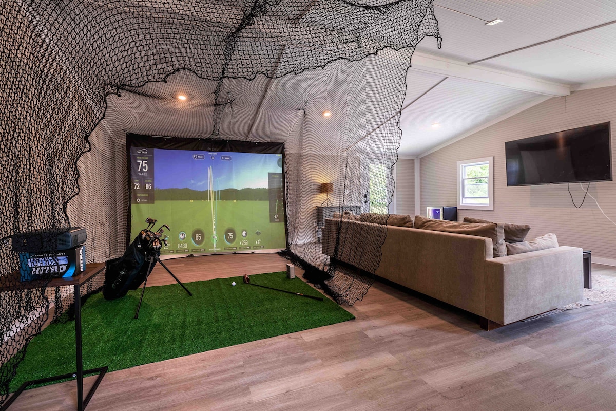 An indoor golf simulator area is equipped with a large projection screen displaying a golf course scene. The artificial turf surface is surrounded by a net for containment. A spacious sectional couch and a mounted TV are visible, providing a comfortable viewing area.
