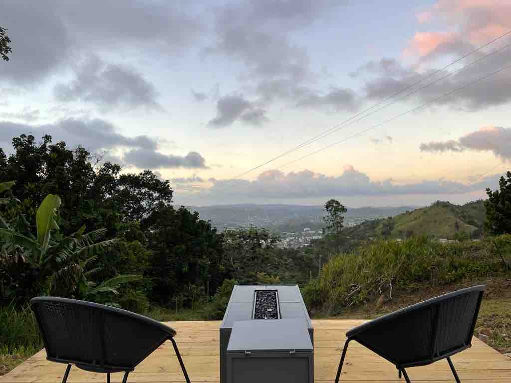 Two black chairs are positioned around a rectangular fire pit on a wooden deck, offering views of the city and rolling hills. The sky displays soft colors of twilight, surrounded by lush greenery and distant mountains.