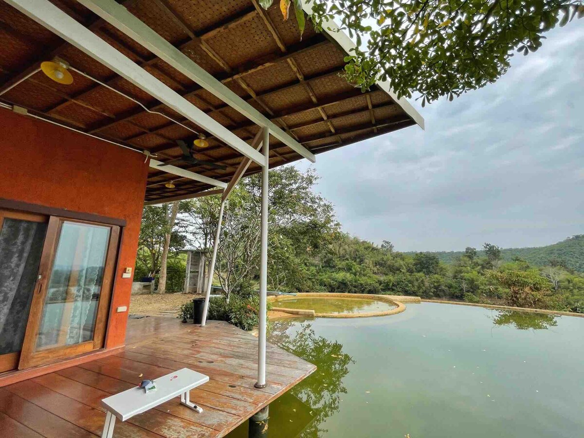 The exterior view showcases a wooden deck extending over tranquil water, surrounded by lush greenery. Large glass doors invite natural light, while a shaded area provides a serene spot to relax. The landscape features gently rolling hills in the background, enhancing the peaceful setting.